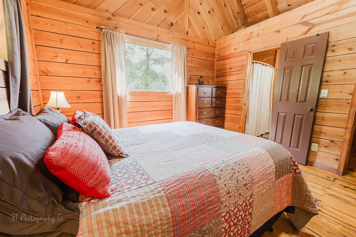A bedroom with a colorful quilted bed, wooden walls, and a window framed by curtains. Natural light spills in, with views that hint at the surrounding nature, connecting the interior comfort to the outdoors.