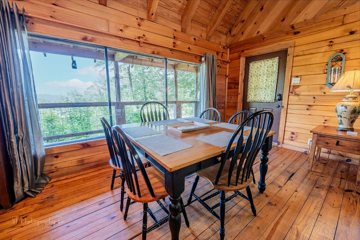 A spacious dining area surrounded by large windows, wooden flooring, and a cozy table illuminated by a warm lamp. The setting is ideal for meals, board games, and conversation, with views that hint at the natural beauty just outside.