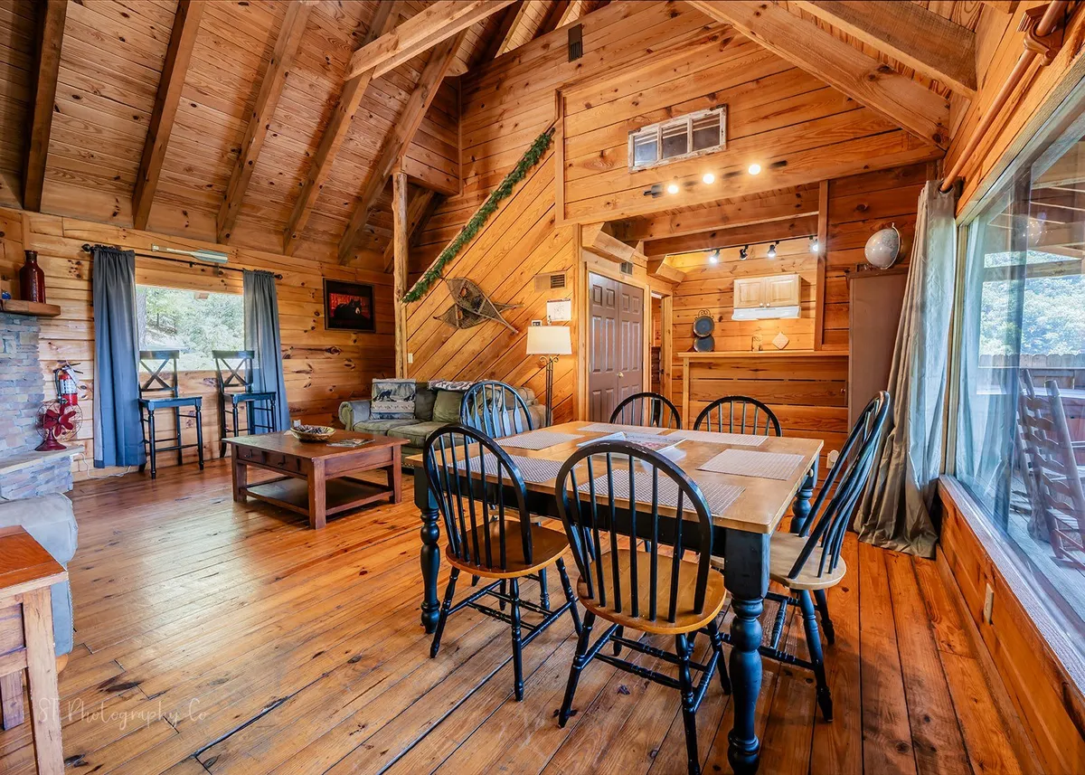 A bright and spacious open-plan interior featuring a dining table with black chairs, a plush sofa area with soft cushions, and expansive windows filling the space with natural light. The setting is ideal for family dining, game nights, or holiday celebrations.