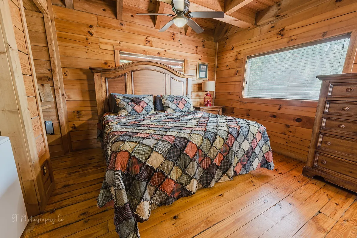 A cozy bedroom featuring a bed with a quilt, wooden furniture, and a window with blinds letting in natural light. The setting is inviting and restful, emphasizing comfort and warmth.