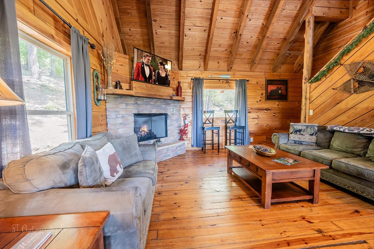A cozy living room with a plush sofa set in front of a stone fireplace, TV mounted above, rustic wooden beams, and a coffee table at the center. The setting is warm, inviting, and perfect for relaxation or social gatherings.