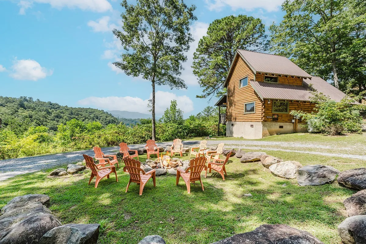 The log cabin exterior is complemented by a cozy fire pit area surrounded by Adirondack chairs. Rolling hills and expansive views provide a stunning backdrop, ideal for gatherings and sunset moments.