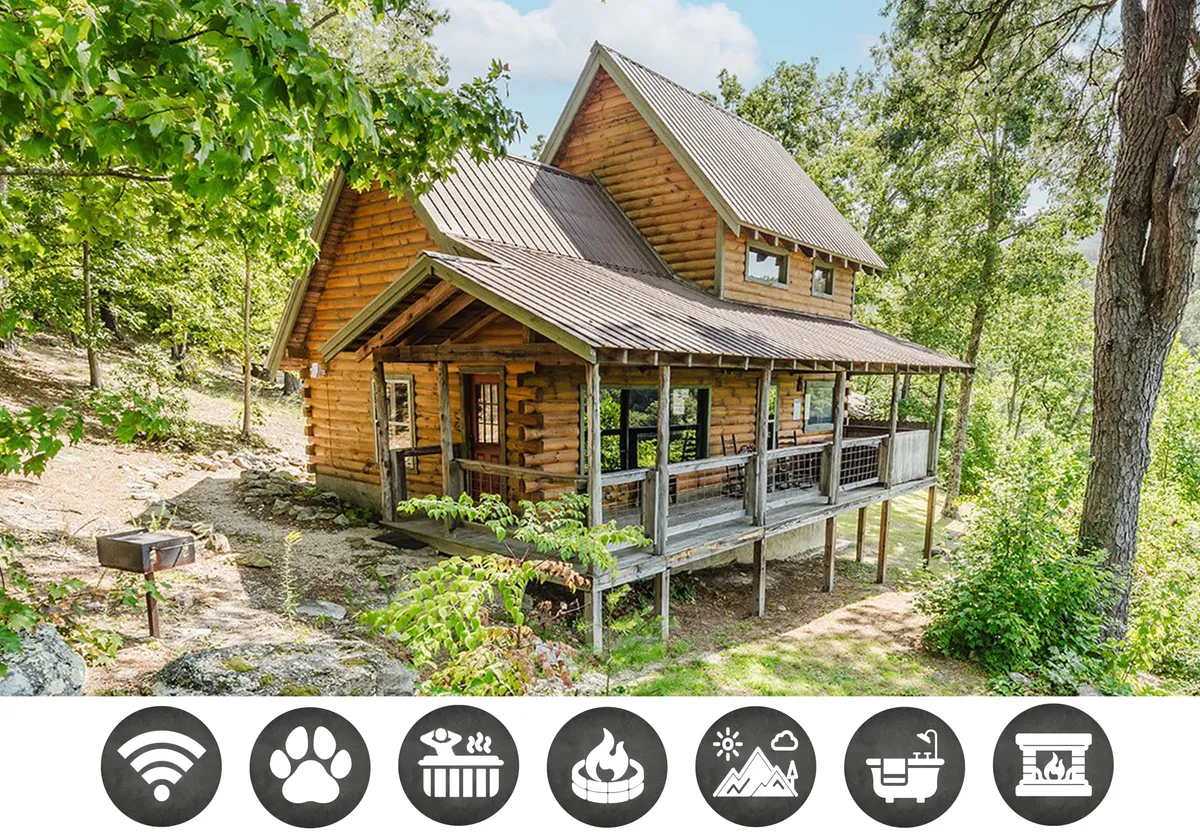 The hero image showcases a charming log cabin surrounded by lush greenery, featuring a spacious wraparound porch perfect for relaxing or enjoying meals outdoors. The setting exudes tranquility and natural beauty, inviting guests to unwind in a peaceful mountain retreat. The porch, visible in the image, hints at moments of sunrise coffee, evening bonfires, and soaking in the mountain air.