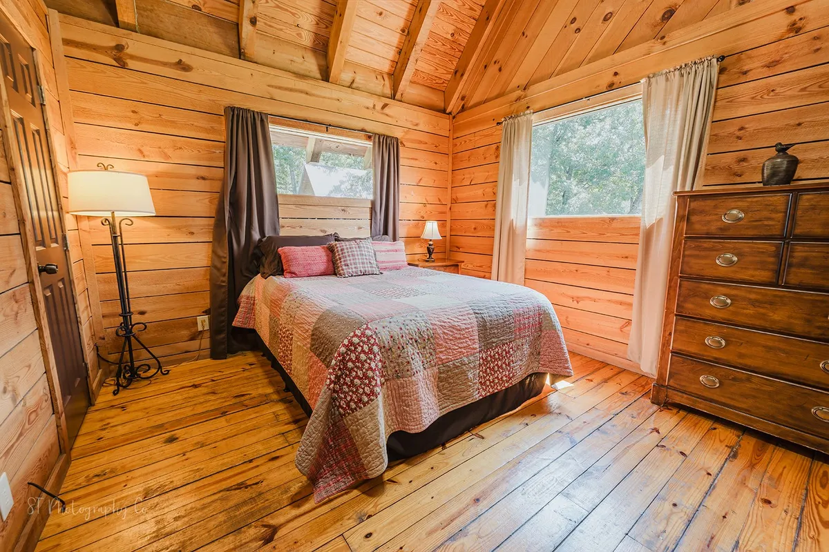 A sunlit bedroom with a patterned quilt on the bed, a wooden dresser, a lamp beside the bed, and large windows letting in natural light. The atmosphere is warm, cozy, and welcoming.