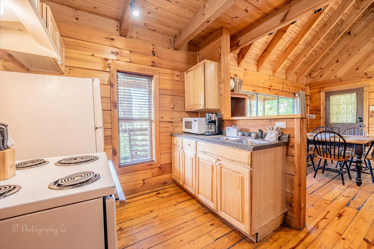 A view of the kitchen area showing a central countertop, wooden cabinetry, windows letting in daylight, and a nearby dining table. The layout is ideal for preparing meals while staying connected with friends and family gathered around the table.