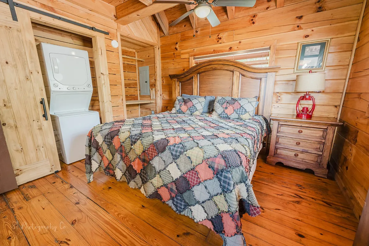 A comfortable bedroom featuring a patterned quilt on the bed, rustic wooden furniture, and a washer-dryer unit subtly integrated into the space. Sunlight streams softly through the windows, creating a warm, lived-in feel.