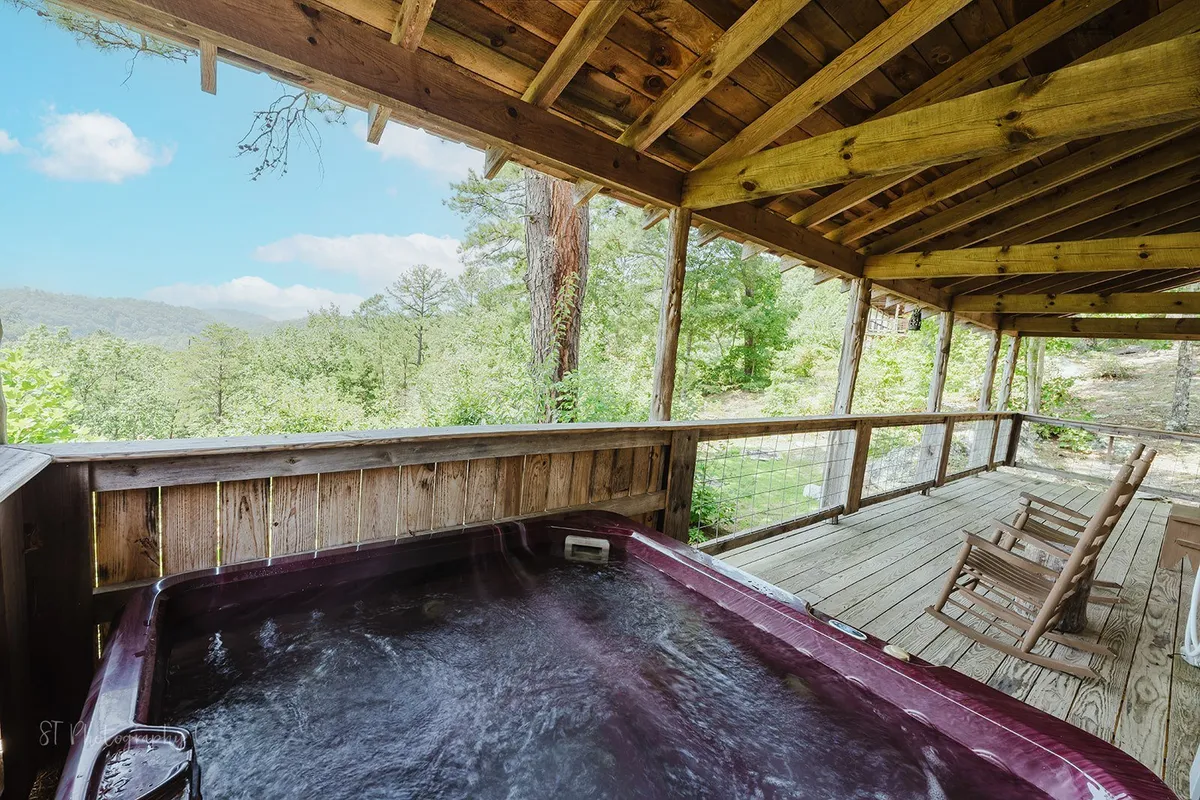 A serene wooden deck nestled among the trees features a bubbling hot tub and classic rocking chairs. Sunlight filters through the foliage, highlighting the inviting space for relaxation and socializing.