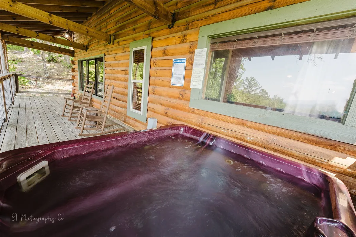 A spacious wooden deck features a hot tub, rocking chairs, and large windows, offering views into both the surrounding nature and the inviting cabin interior. The setup is ideal for socializing, relaxing, and enjoying scenic vistas as a family.