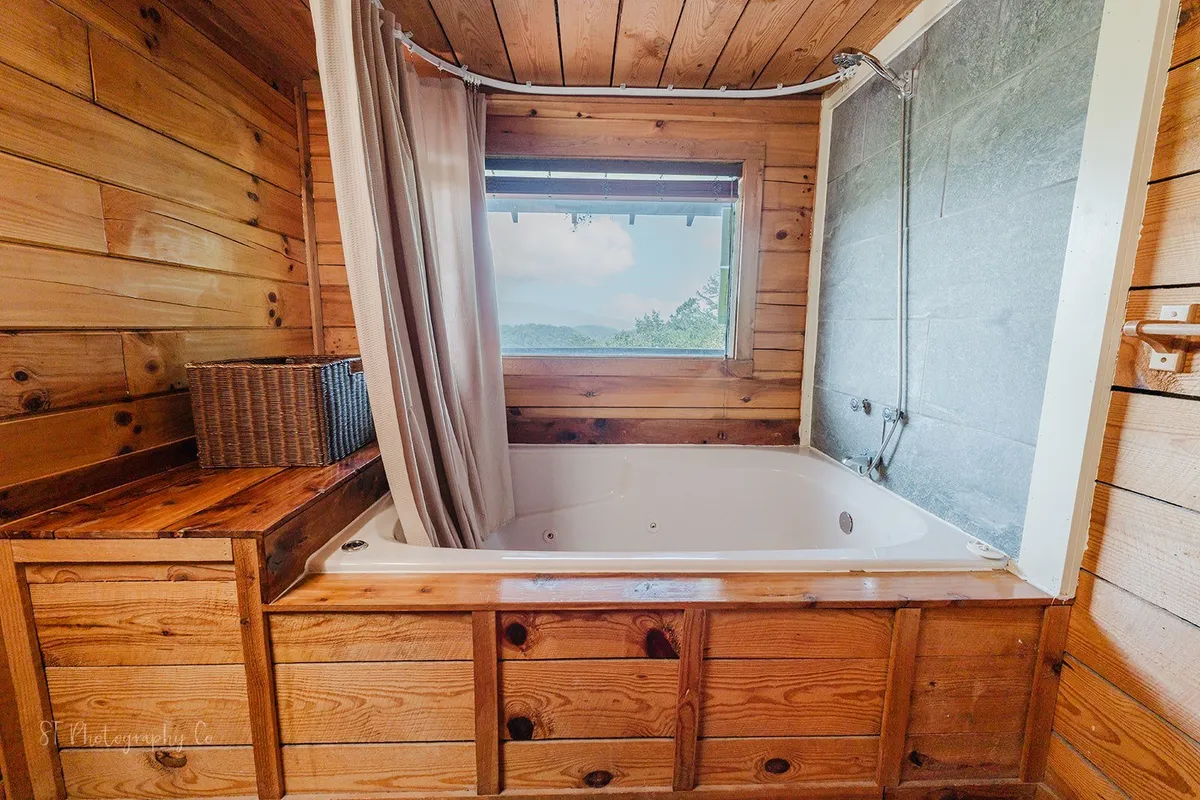 A bright, tranquil bathroom with a whirlpool tub positioned under a window offering scenic views. Wooden cabinetry enhances the cozy ambiance, making the space perfect for relaxation and rejuvenation.
