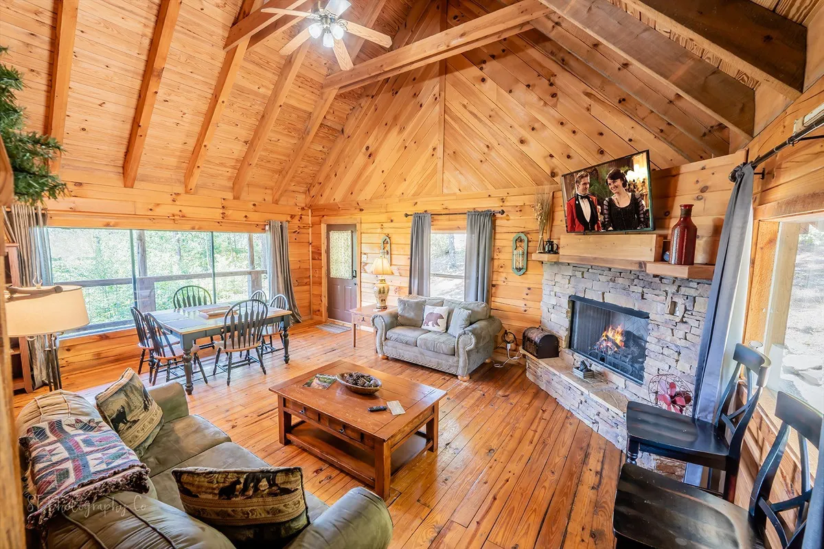 A warm and inviting living room featuring a stunning stone gas fireplace, plush sofas, large windows letting in natural sunlight, and exposed wooden beams. The arrangement of furniture encourages cozy gatherings and relaxation, with a perfect spot to unwind or play board games by the fire.