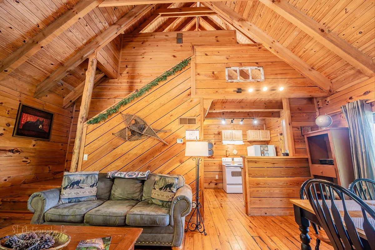 An open view of the living space where a comfortable sofa with decorative pillows sits under wooden beams, facing a spacious kitchen and a dining table. The area is filled with natural light from nearby windows, creating a seamless flow between lounging, cooking, and dining spaces.
