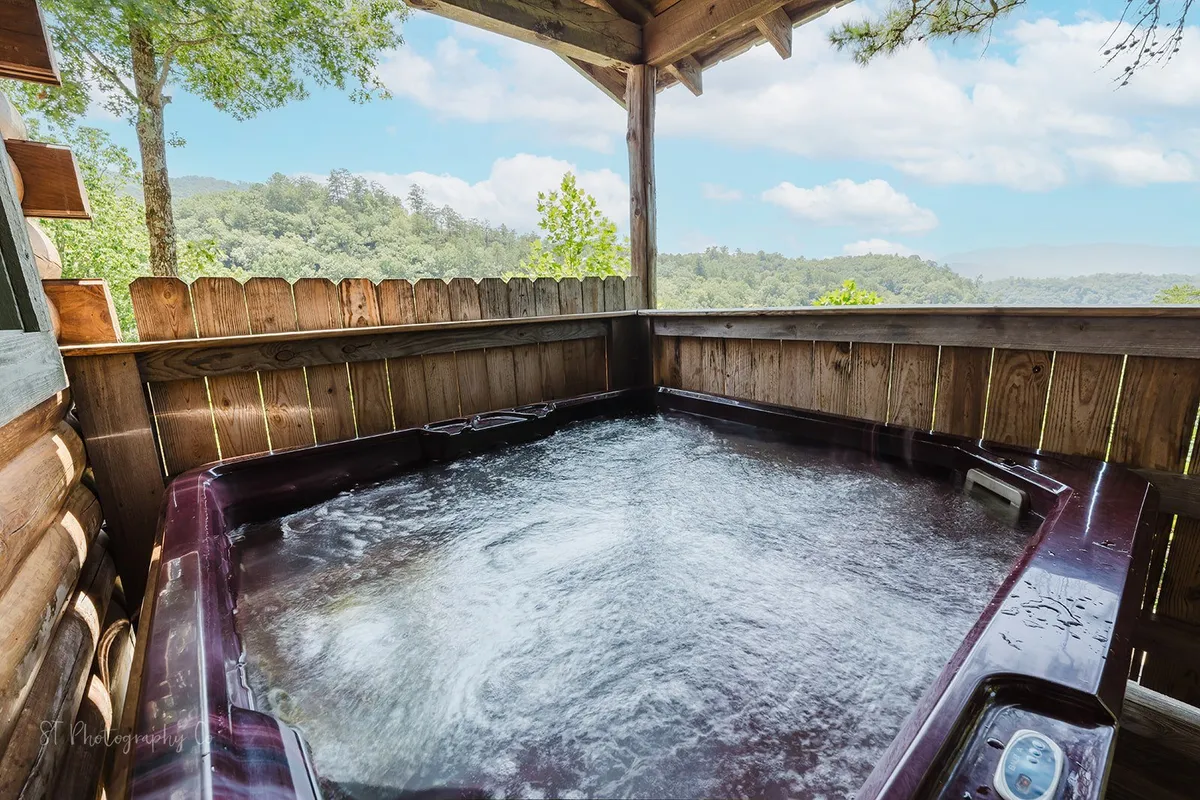 An inviting hot tub sits on a wooden deck, surrounded by a scenic view of the lush outdoors and a wooden fence. The peaceful setting evokes relaxation after a day of exploration, highlighting the cabin's tranquil amenities.