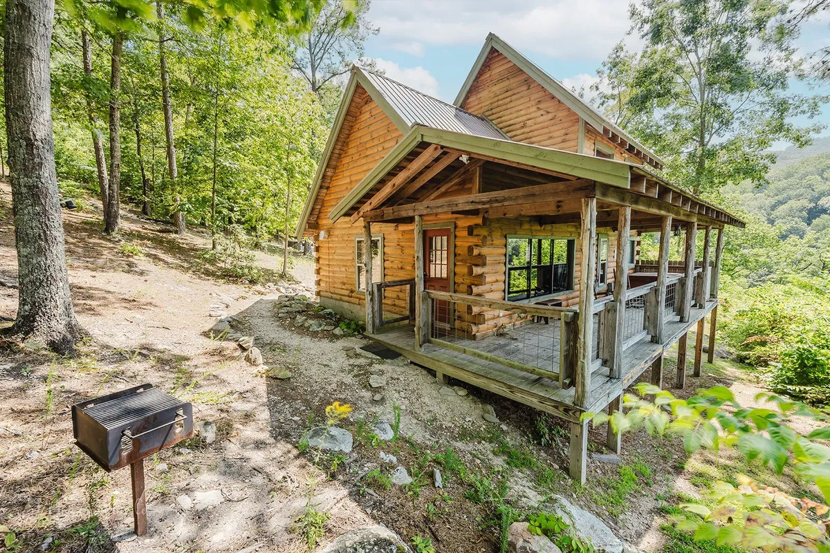 A classic log cabin with a wrap-around porch and an outdoor grill set amidst lush greenery. The inviting porch hints at opportunities for leisure, while the grill signals alfresco dining possibilities.